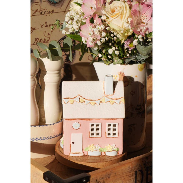 Der Isabelle Rose - Keramik Haus als Teelichthalter im Landhausstil Rosa, ein rosafarbener Teelichthalter in Form eines Hauses aus Keramik, steht auf einem Holztablett neben einem weißen Krug mit rosa und weißen Blumen.