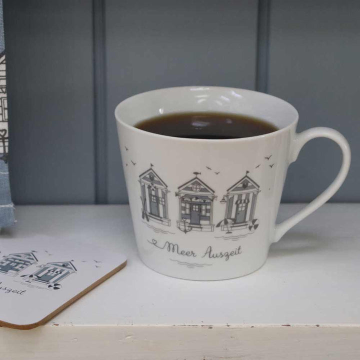 Ein Krasilnikoff - Happy Cup Meer Auszeit mit schwarzen Illustrationen von Strandhäusern und dem Text "Meer Auszeit" enthält eine dunkle Flüssigkeit. Ein passender Untersetzer ist links teilweise zu sehen.