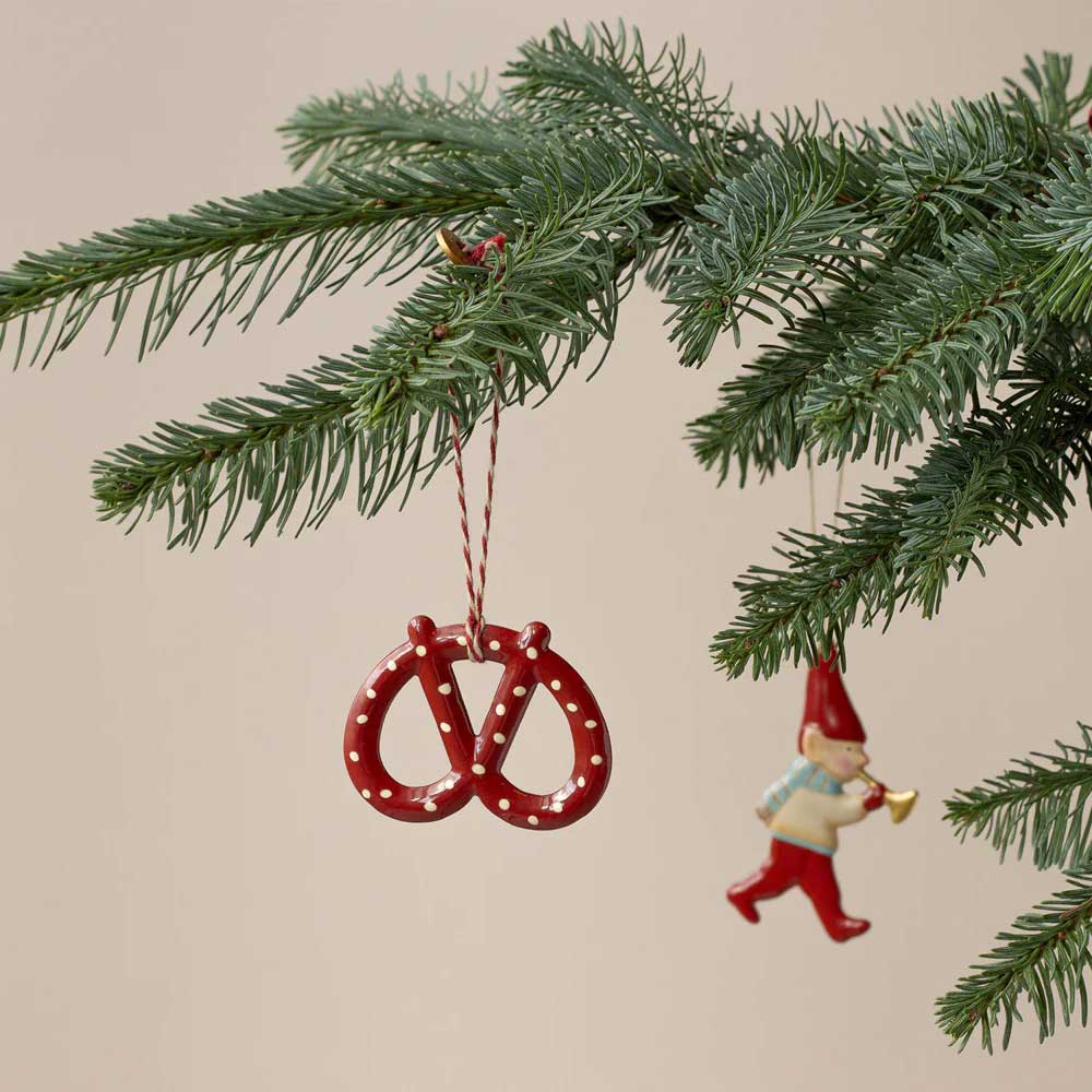 Ein Weihnachtsbaumzweig mit zwei hängenden Ornamenten: Das eine ist der rote Maileg-Anhänger Brezel Metall und das andere ist eine kleine Figur eines Gnoms, der Trompete spielt.