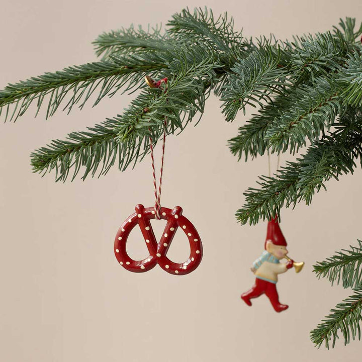 Ein Weihnachtsbaumzweig mit zwei hängenden Ornamenten: Das eine ist der rote Maileg-Anhänger Brezel Metall und das andere ist eine kleine Figur eines Gnoms, der Trompete spielt.