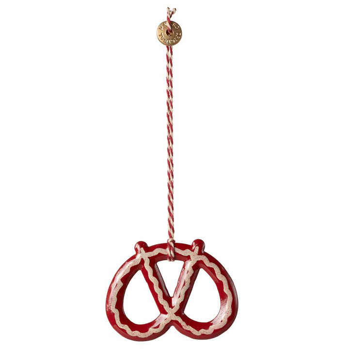 Der Maileg - Anhänger Brezel Metall ist ein brezelförmiges Ornament in Rot und Weiß, das an einer dekorativen Schnur hängt und an dem ein kleines rundes Etikett befestigt ist.