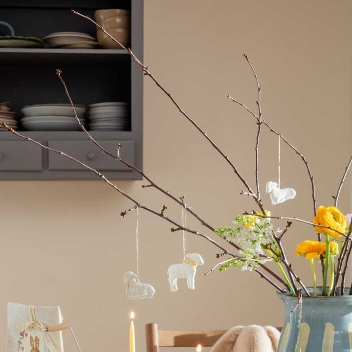 Eine ruhige Esszimmerecke mit einer Vase mit gelben Blumen und zarten hängenden Maileg - Anhänger Lamm Metall-Ornamenten, brennenden Kerzen und einem offenen Regal mit Geschirr.