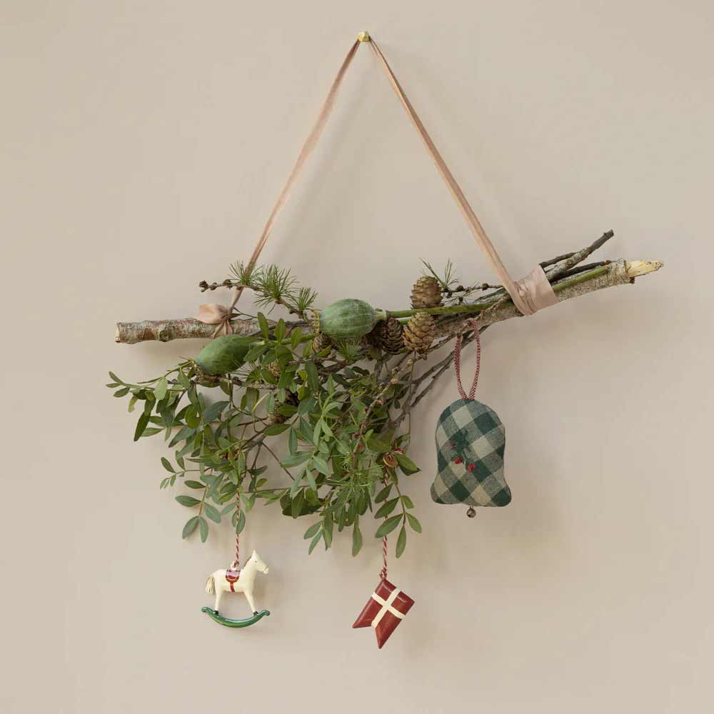 An einer Wand hängt ein Zweig mit grünen Blättern und Tannenzapfen, geschmückt mit einem Stoffglöckchen, dem Maileg - Anhänger Metall Schaukelpferd und einem rot-weißen Flaggenornament.