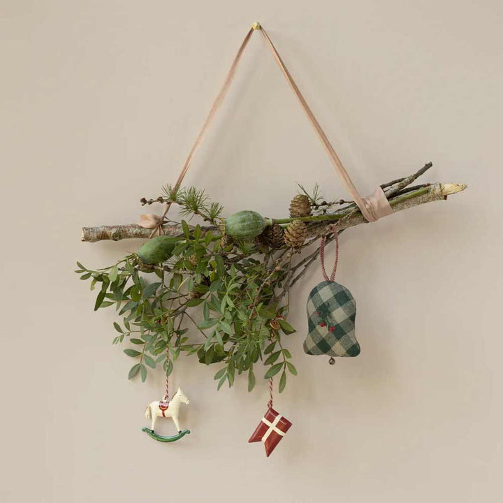 An einer Wand hängt ein Zweig mit grünen Blättern und Tannenzapfen, geschmückt mit einem Stoffglöckchen, dem Maileg - Anhänger Metall Schaukelpferd und einem rot-weißen Flaggenornament.