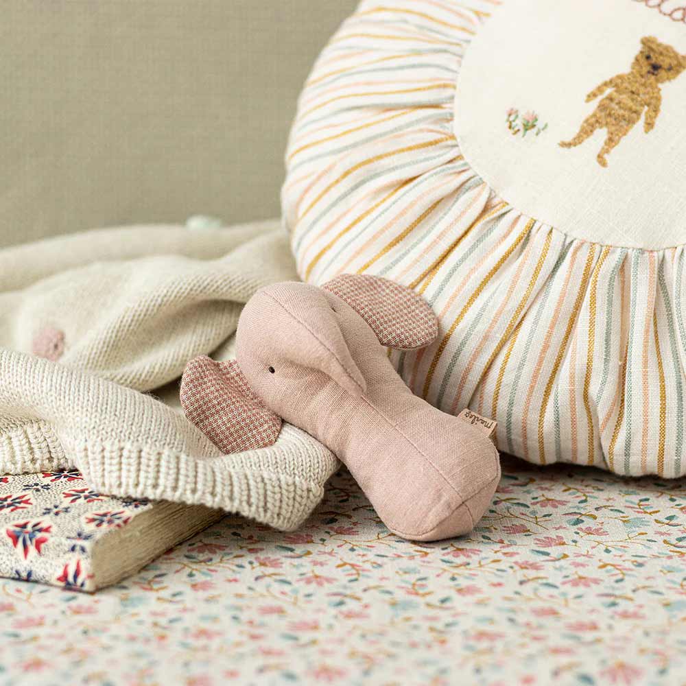 Ein Maileg - Elefant Rassel Lullaby Friends liegt auf einer gemusterten Oberfläche neben einem runden Kissen mit einer Bärenillustration, einem Strickkleidungsstück und einem Buch mit Blumeneinband.