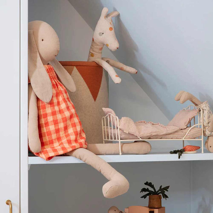 Ein Maileg-Hase im Kleid Größe 5 (Höhe: 64 cm) in einem orangefarbenen Kleid sitzt mit einem anderen kleinen Stofftier auf einem Regal neben einem Spielzeugbett.