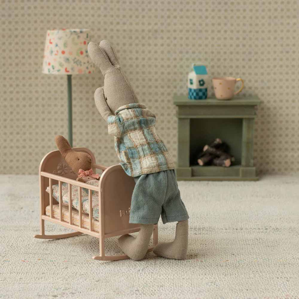 Der Maileg - Hase in Hemd und Shorts Größe 1 kniet neben einer Krippe, in der sich ein weiterer Hase befindet; eine Spielzeuglampe, ein Kamin und eine Tasse schmücken das Miniaturzimmer.