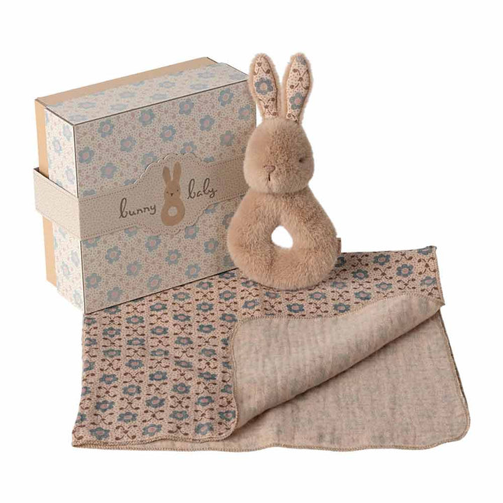 Das Maileg - Hase Rassel-Set Creme Pfirsich ist ein Geschenkset mit einer gemusterten Schachtel, einer plüschigen Hasenrassel und einer dazu passenden gestrickten Babydecke in Beige- und Blautönen.