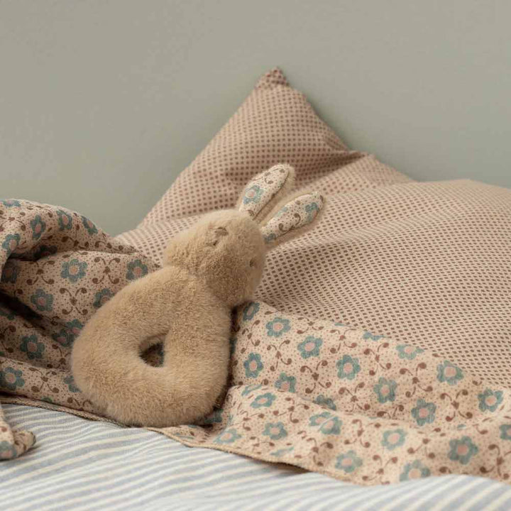 Das Plüschhäschen Maileg - Hase Rassel-Set Creme Pfirsich liegt auf einem beige gemusterten Kissen und einer Decke auf einem ordentlich gemachten Bett mit gestreiften Laken.