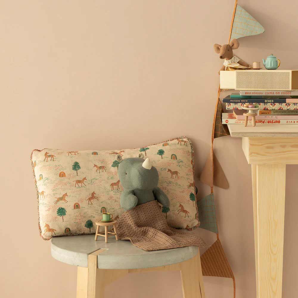 Ein kleiner Hocker mit einem Maileg - Kissen Einhorn, einem grauen Plüschnashorn und einem braunen Tuch steht neben einem Holzregal mit Büchern, einer Spielzeugmaus und einer blauen Teekanne.