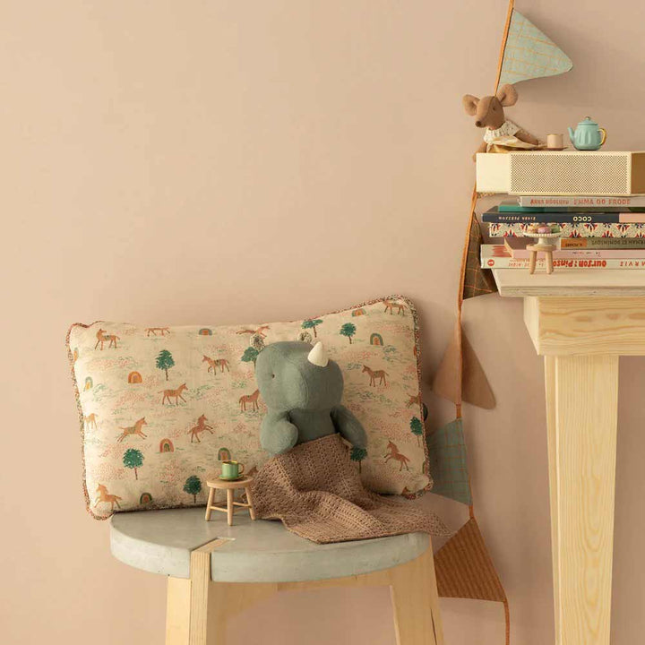 Ein kleiner Hocker mit einem Maileg - Kissen Einhorn, einem grauen Plüschnashorn und einem braunen Tuch steht neben einem Holzregal mit Büchern, einer Spielzeugmaus und einer blauen Teekanne.
