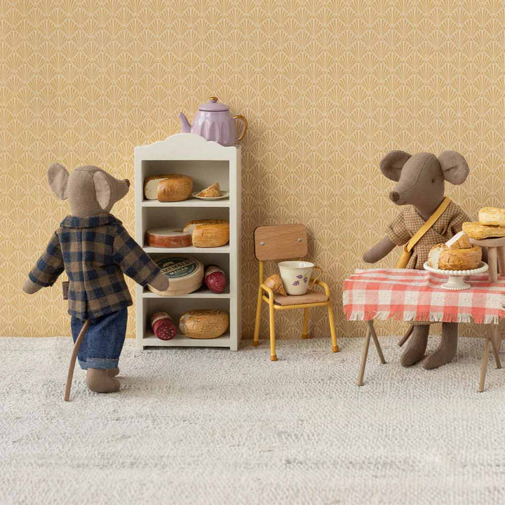 Zwei Mäuse in einer Küche: Eine steht neben einem Brotregal, die andere sitzt mit einer Teetasse an einem mit Gebäck beladenen Tisch. Die Wand ist gemustert, und auf dem Regal steht eine Teekanne. Die stehende Maus trägt Maileg - Maus Kleidung Braunes kariertes Hemd & Hose Papa.