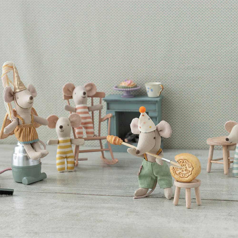 Die Maileg-Figuren „Maus Kleiner Bruder“ Clownskleidung im Koffer sind in einer verspielten Szene dargestellt: Eine Maus sitzt und backt, eine andere saugt Staub und zwei Mäuse entspannen auf Stühlen vor einem Minitisch mit Desserts.