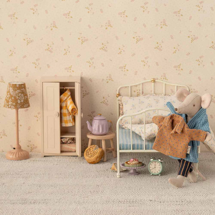 Eine Maileg-Mausfigur steht neben einem Miniaturbett und hält ihre „Maus Puppenkleidung Hemd und Hose Papa“, umgeben von einer kleinen Lampe, einem Kleiderschrank, einer Teekanne, einer Tasse, Gebäck und einer Uhr vor dem Hintergrund einer geblümten Tapete.