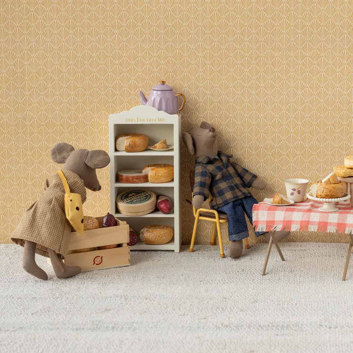 Zwei ausgestopfte Mäusefiguren stehen neben einer Puppenhaus-Bäckerei mit Gebäck, einem mit Essen bedeckten Tisch und einer Kiste. Eine Maus trägt ein Maileg Maus Puppenkleidung Kleid und Tasche Mama-Outfit, die andere ein kariertes Hemd.