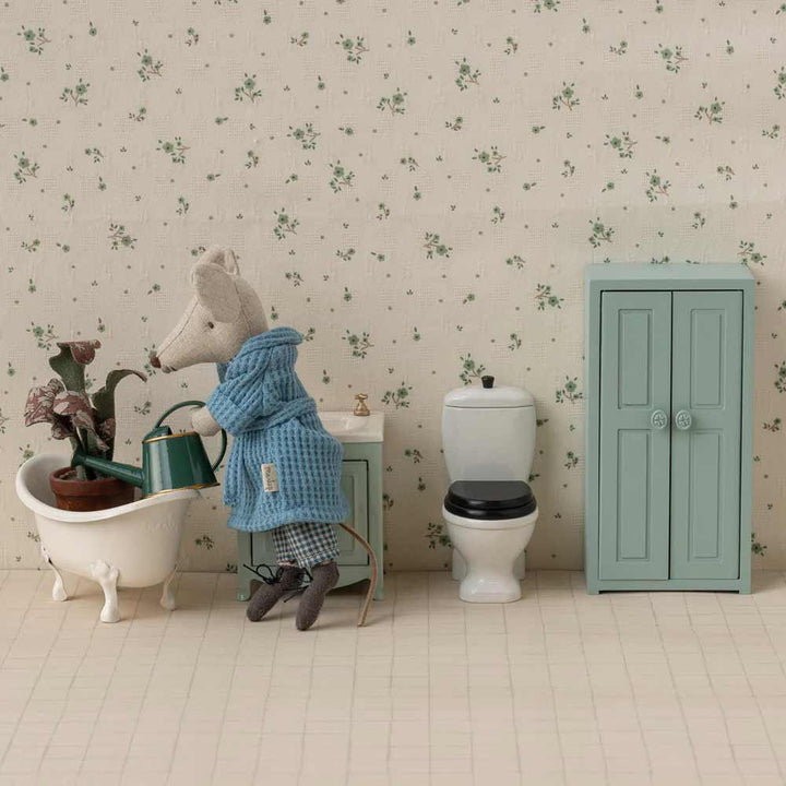 Eine Maileg Miniatur-Gießkanne für Maus wird von einer Spielzeugmaus im blauen Gewand verwendet, um eine Pflanze in einer winzigen Badewanne neben einer Toilette, einem Waschbecken und einem grünen Schrank vor einer geblümten Tapete zu gießen.