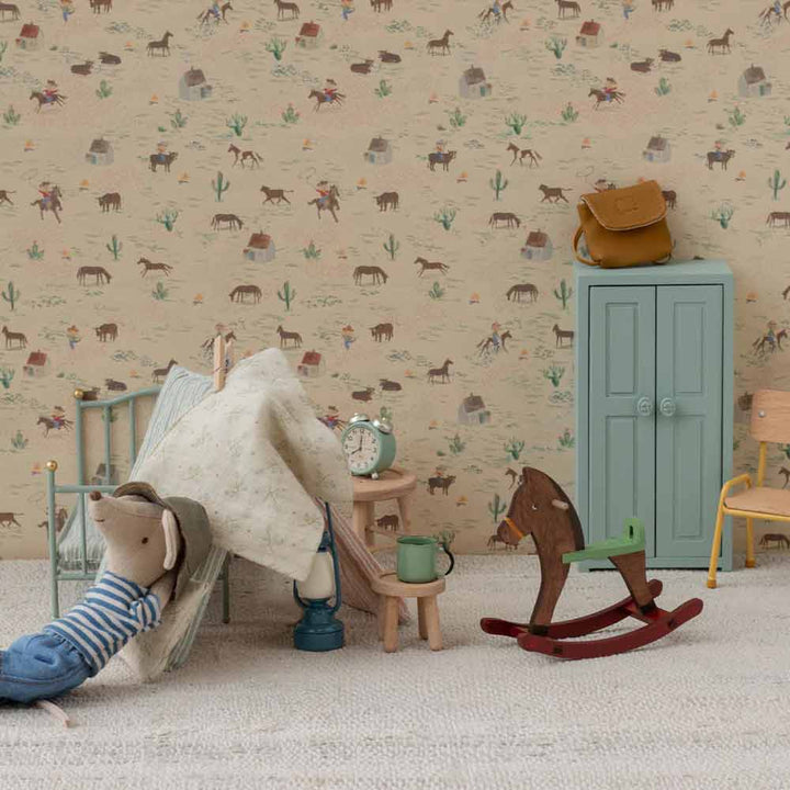 Ein kleines Zimmer mit Spielzeugmöbeln, einem Maileg Miniatur Schaukelpferd für Maus Dunkelbraun, einer Spielzeugmaus im Bett, einem Schrank, Stühlen, einer Spielzeuguhr und einer Tapete mit Tiermotiven.