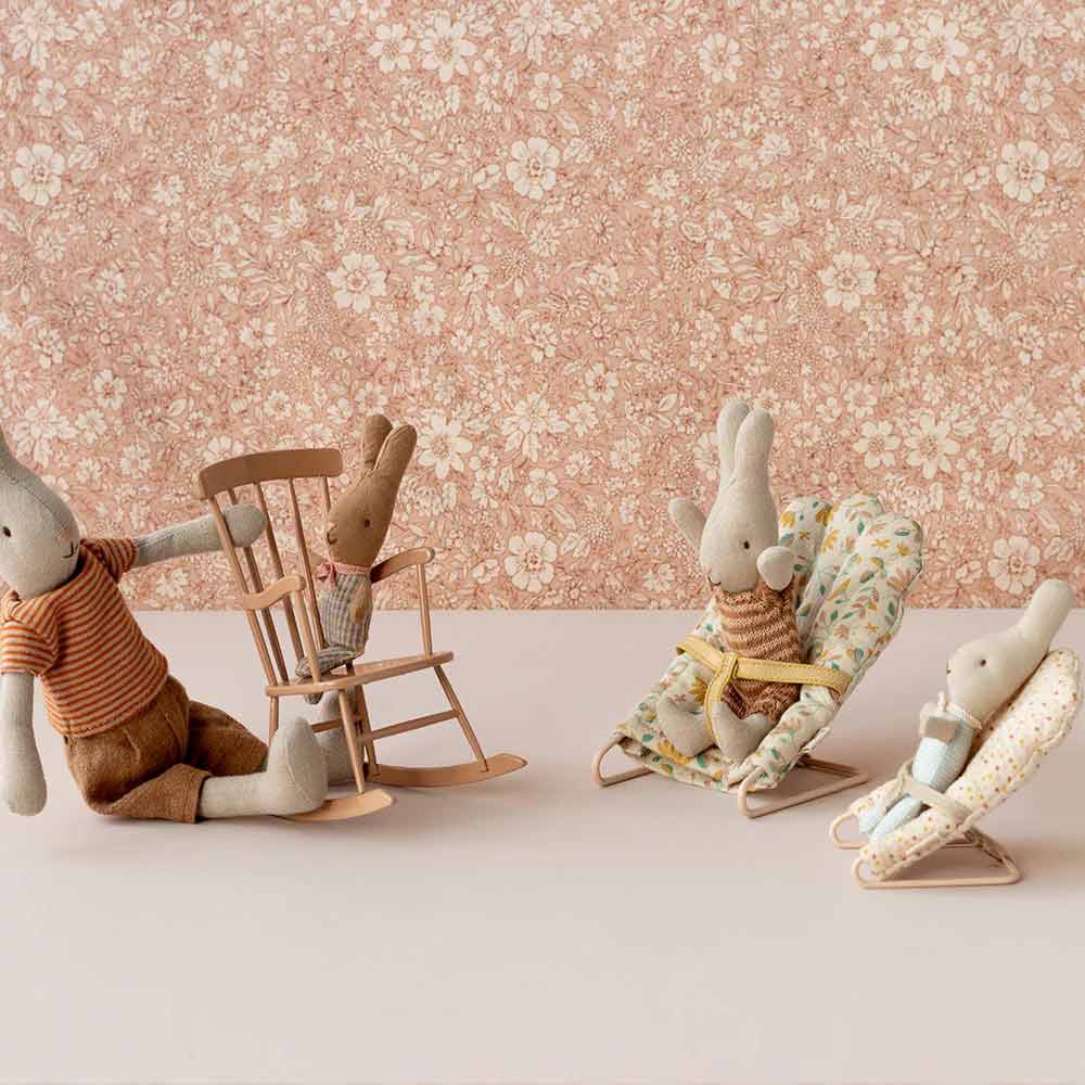 Vier Stoffhasenpuppen stehen in einem Raum mit Blumentapete. Ein erwachsener Hase sitzt mit einem Baby in einem Schaukelstuhl, und zwei weitere Hasen sitzen in Maileg-Babysitter-Mikro-Stühlen.