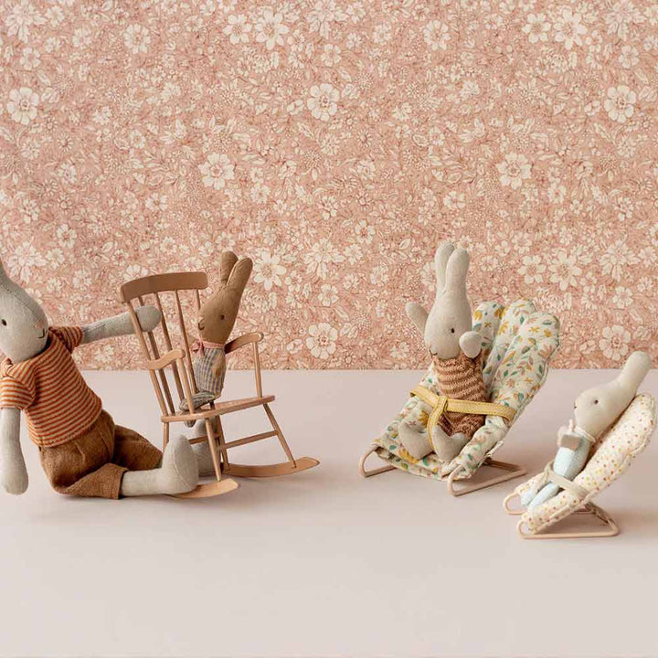 Vier Stoffhasenpuppen stehen in einem Raum mit Blumentapete. Ein erwachsener Hase sitzt mit einem Baby in einem Schaukelstuhl, und zwei weitere Hasen sitzen in Maileg-Babysitter-Mikro-Stühlen.