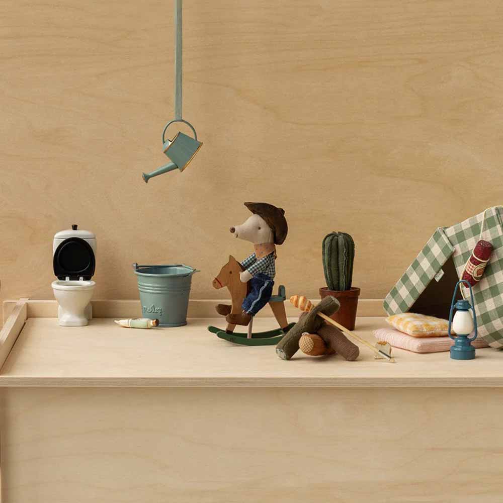 Der kleine Bruder der Maileg - Maus Cowboy auf Schaukelpferd sitzt zwischen winzigen Campinggegenständen wie einem Zelt, einem Kaktus, einem Eimer, einer Gießkanne, einer Laterne, einer Toilette, Brennholz und einem Kissen auf einer Holzoberfläche.