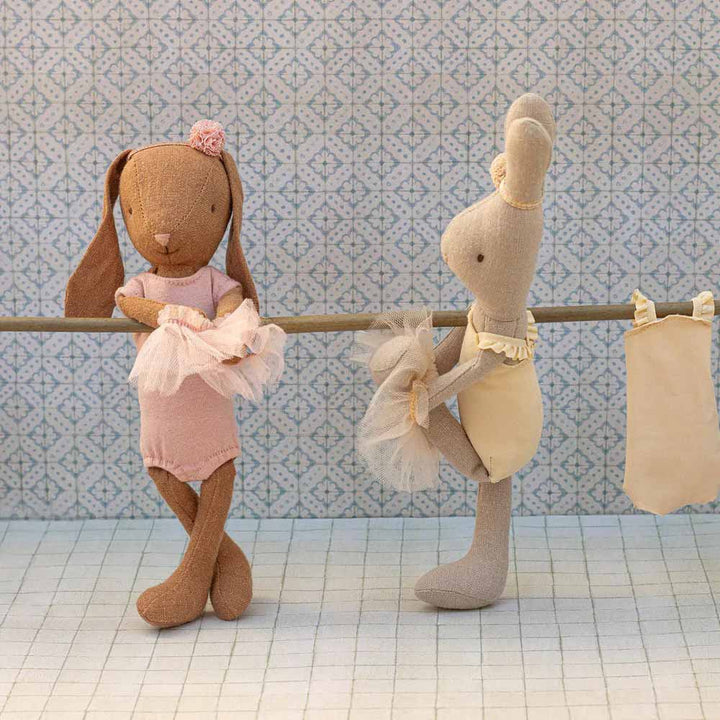 Zwei Maileg-Hasenpuppen tragen die Outfits Puppenkleidung Hase Ballettanzug und Rock Größe 1 und stehen neben einer Stange, an der ein Tutu vor einem gemusterten Hintergrund hängt.