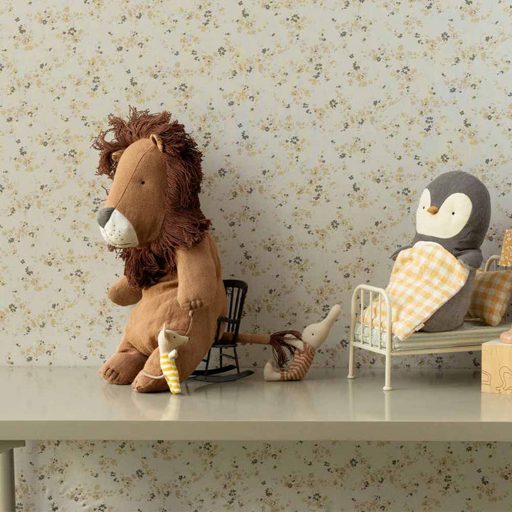 Maileg Safari Friends Löwe Medium, zusammen mit Mäusen und Pinguinspielzeug, arrangiert auf einem Tisch vor einem Hintergrund aus Blumentapeten, wobei der Löwe auf einem Stuhl sitzt und der Pinguin in einem Papierbett liegt.