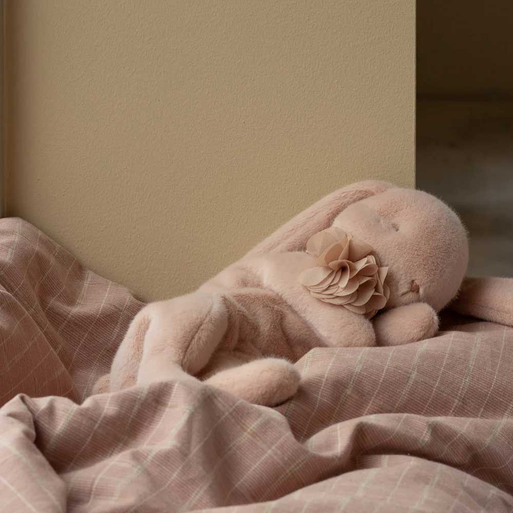 Der Maileg - Schlafender Plüsch Hase, ein Plüschhase mit Schlappohren und Blumendetail, ruht auf rosa kariertem Stoff neben einer beigen Wand.