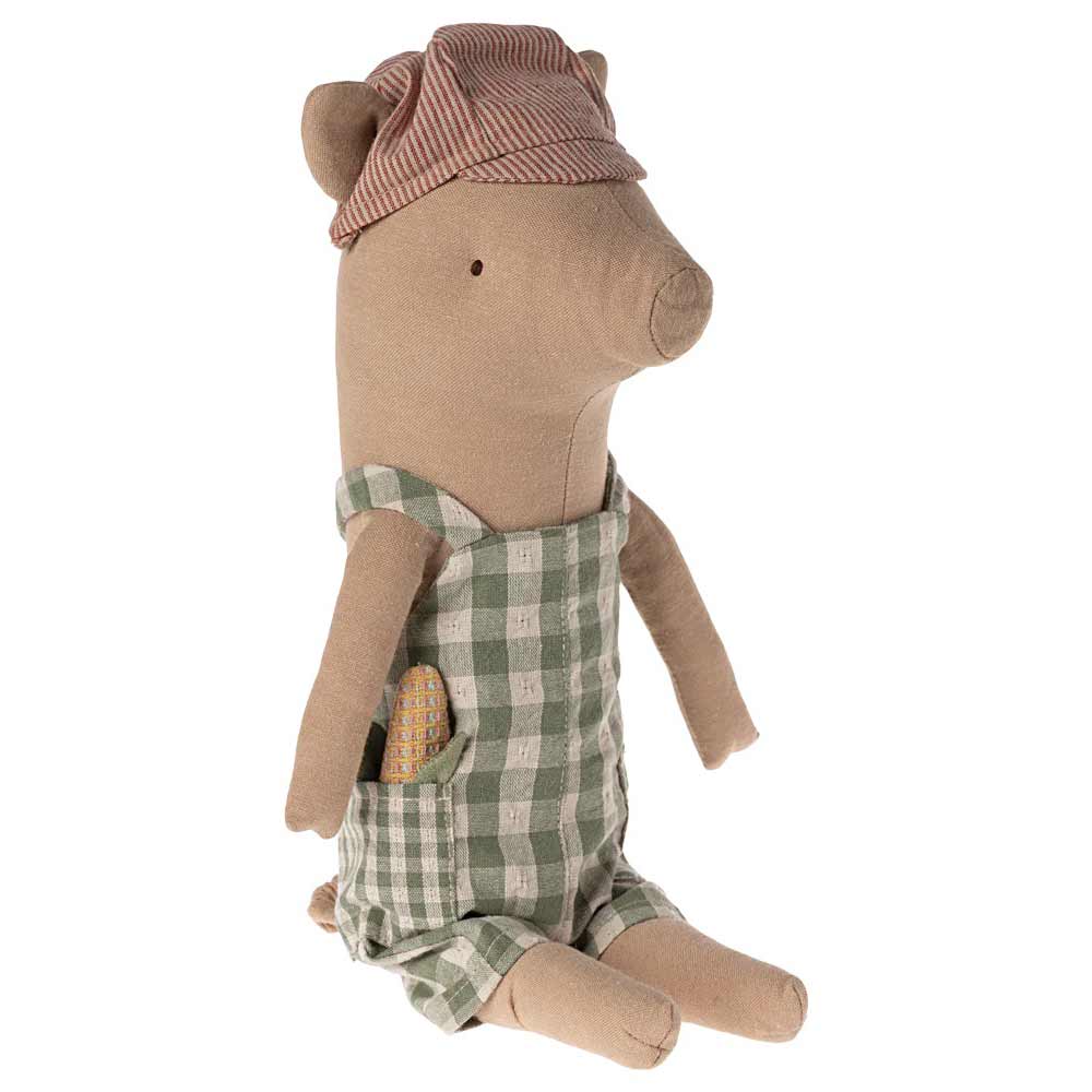 Ein Maileg - Schwein Junge, bekleidet mit einem grün karierten Overall und einer rot gestreiften Mütze, sitzt mit einem kleinen Gegenstand in der Tasche da.