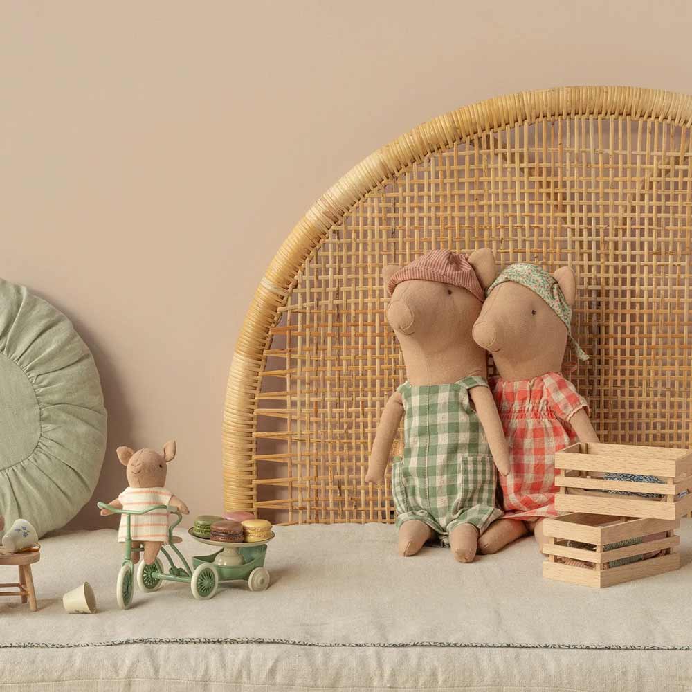 Zwei Stoffmauspuppen in karierter Kleidung sitzen auf einem Kopfteil aus geflochtenem Rattan. Ein kleineres Maileg-Schwein Junge auf einem Dreirad und Holzkisten stehen in der Nähe auf einer Stoffoberfläche.