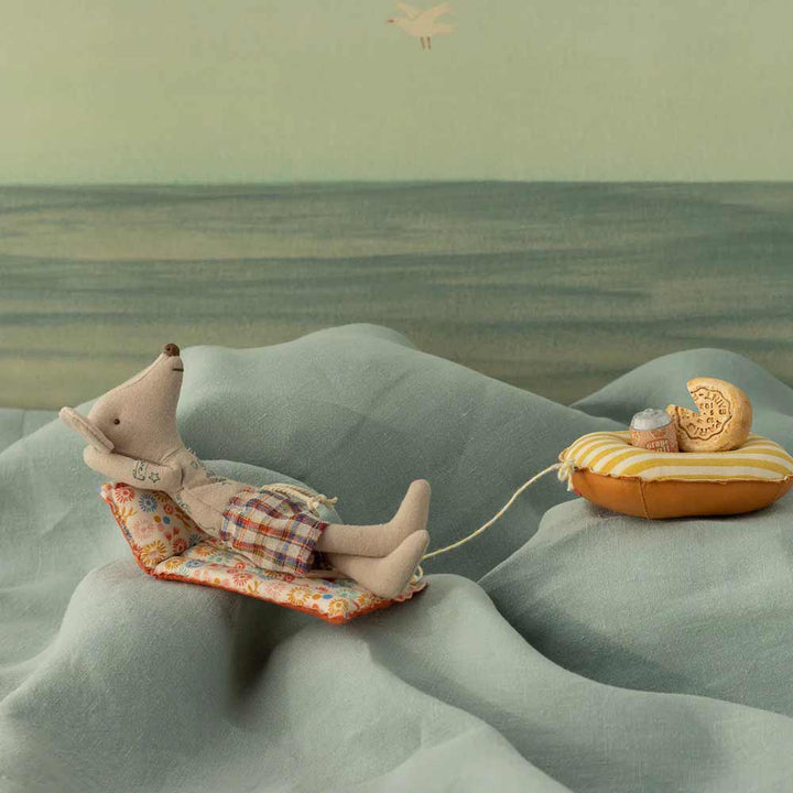 Der Maileg - Strandmaus Papa mit Luftmatratze zeigt einen entspannten Mäusepapa auf einem gemusterten Liegestuhl, der mit einer Schnur an einem Schwimmkörper und einem Spielzeugboot mit einer winzigen Maus und Käse befestigt ist, alles eingebettet in eine bezaubernde Stofflandschaft.