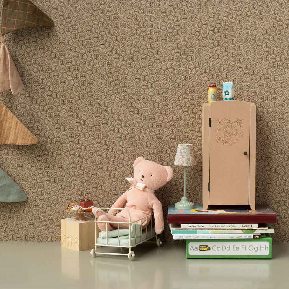 Das Maileg - Teddy Rose sitzt in einer Spielzeugkrippe auf gestapelten Büchern neben einem kleinen Schrank, einer Lampe und einem Wimpel vor einer gemusterten Tapete.
