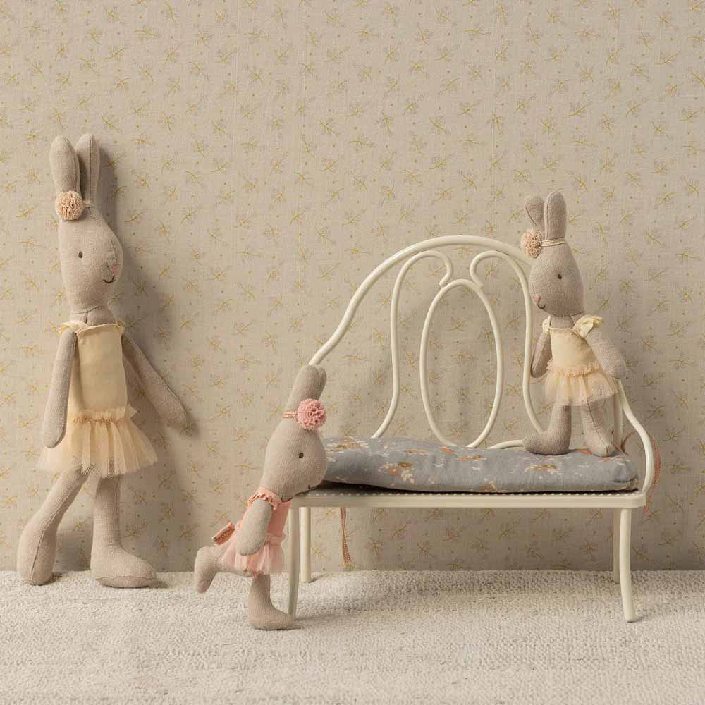Drei Stoffhasenpuppen in pastellfarbenen Kleidern posieren um die Maileg Vintage Bank Miniatur Off White Bank mit gepolsterter Sitzfläche vor einer gemusterten Wand.
