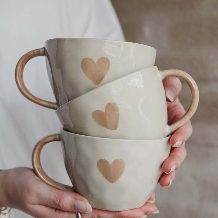 Eine Person in einem weißen Hemd hält drei stapelbare Majas Cottage-Becher, jeder davon mit Herzmotiven verziert, mit der Aufschrift „Eine schöne Tasse Liebe und Glück“.