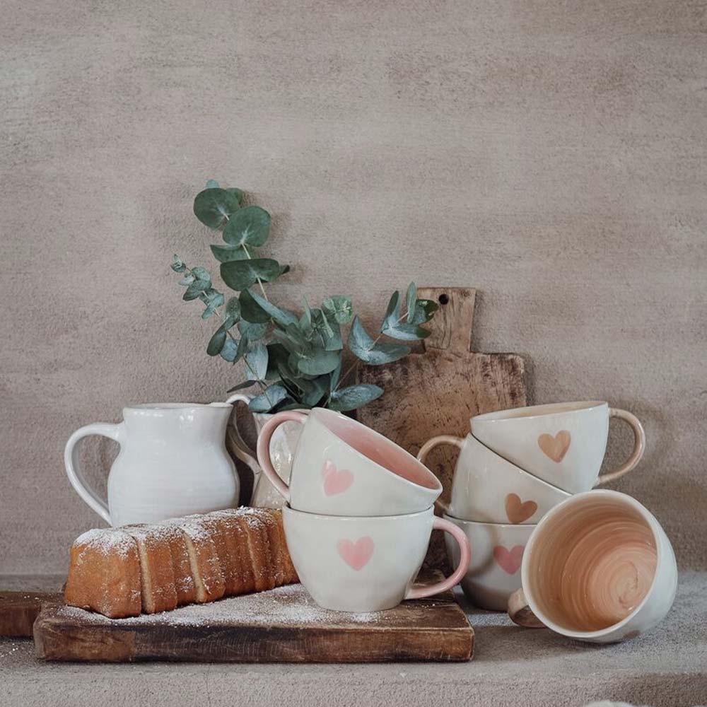 Majas Cottage – Eine schöne Tasse „Love & Happiness“ mit Herzmotiven ist neben einem kleinen weißen Krug, einem mit Zucker bestäubten Brotlaib und Eukalyptuszweigen auf einem Holzbrett gestapelt.