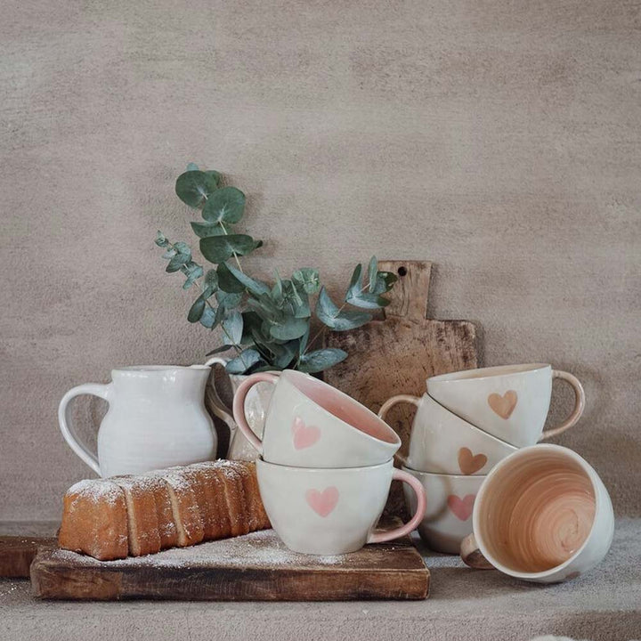 Majas Cottage – Eine schöne Tasse „Love & Happiness“ mit Herzmotiven ist neben einem kleinen weißen Krug, einem mit Zucker bestäubten Brotlaib und Eukalyptuszweigen auf einem Holzbrett gestapelt.