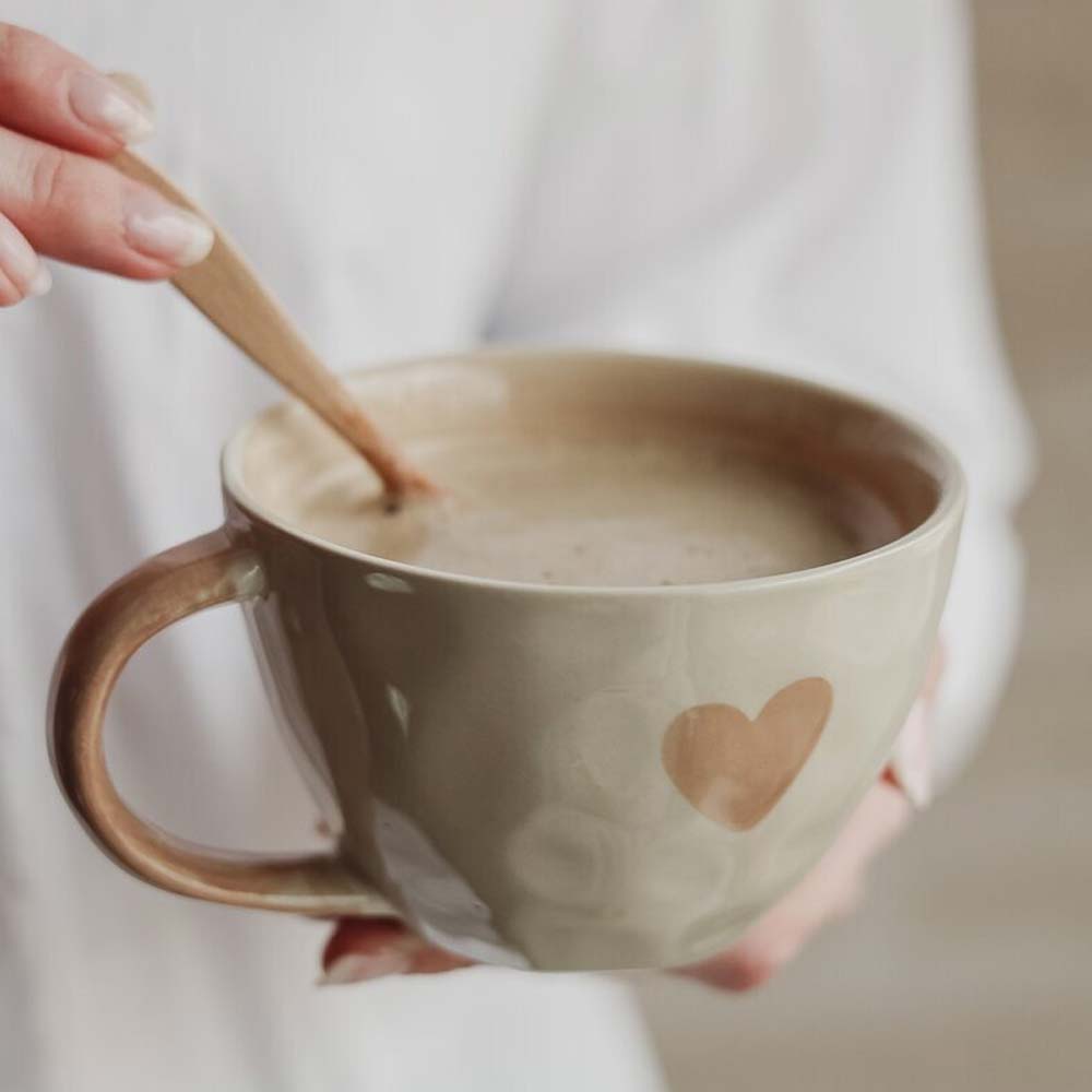 Im Majas Cottage „A lovely cup of Love & Happiness“, einem großen beigen Becher mit Herzmuster, rührt eine Person mit einem Holzlöffel einen Latte um.