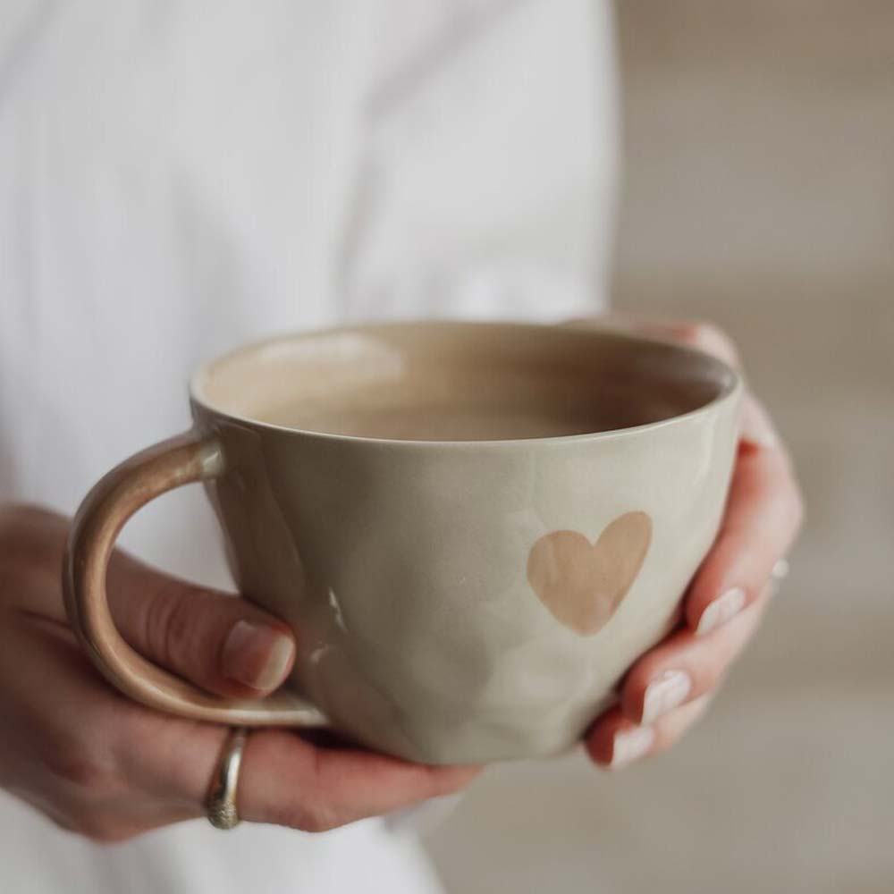 Jemand hält eine Majas Cottage „Eine schöne Tasse Liebe und Glück“, beige mit Herzdesign, gefüllt mit einem heißen Getränk.