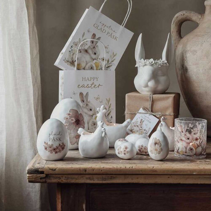 „Deko Ei mit Blume“ von Majas Cottage sorgt zusammen mit Keramikeiern, Hasen und Hühnern mit floralen Motiven für eine festliche Osterszene auf einem Holztisch, im Hintergrund mit Geschenktüten und Geschenken geschmückt.