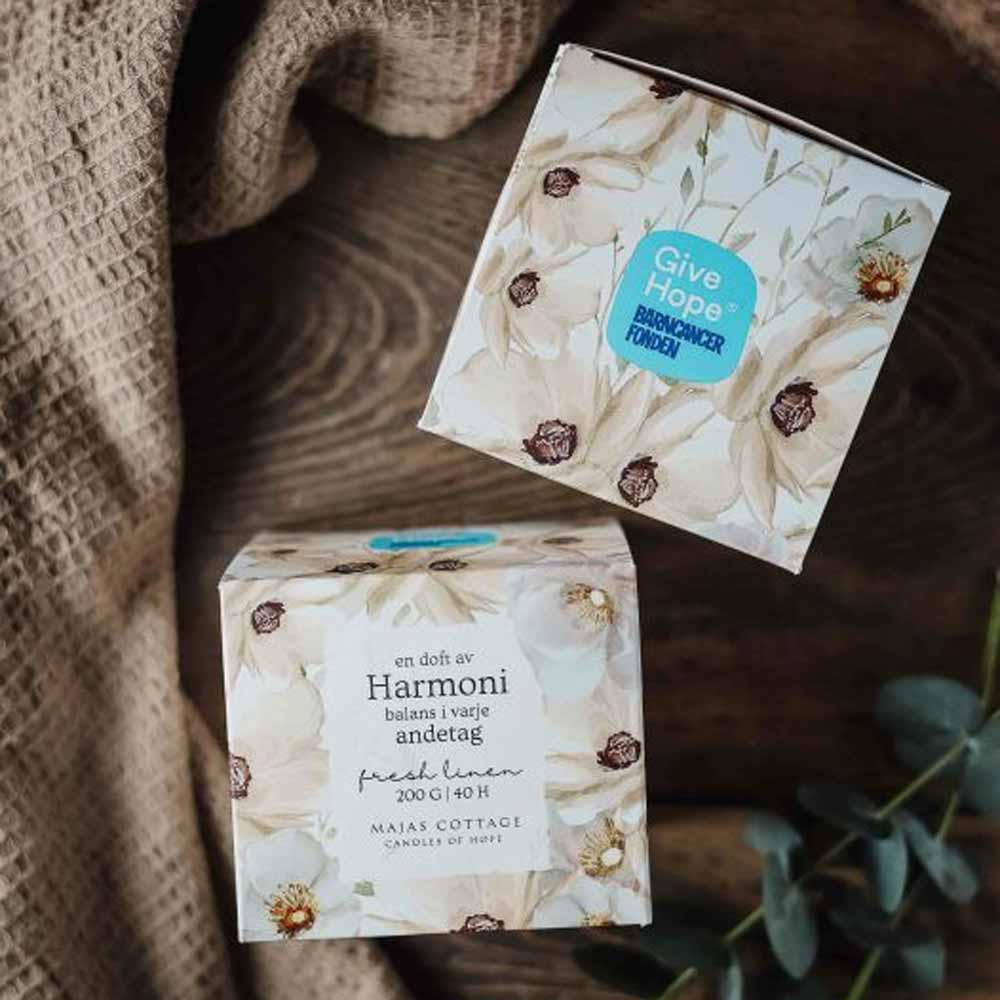 Zwei dekorative Schachteln mit floralen Mustern auf einer Holzoberfläche, eine mit der Aufschrift „Harmoni“ und die andere mit einer Duftkerze namens „Majas Cottage – Duftkerze Frischer Leinen mit Geschenkebox 200g“.