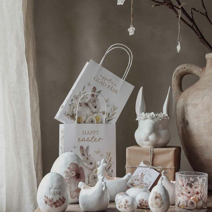 Oster-Setup mit „Frohe Ostern“-Geschenktüten von Majas Cottage, Hasen- und Hühnerfiguren aus Keramik, bemalten Eiern und einem verpackten Geschenk, alles auf einem Tisch neben einer großen Vase arrangiert.