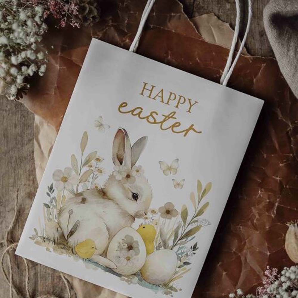 Die Majas Cottage - Geschenketüte Happy Easter zeigt einen Hasen, Blumen, bemalte Eier und ein Küken mit dem Aufdruck „Frohe Ostern“. Sie ist auf braunem Papier platziert und von kleinen weißen Blumen umgeben.