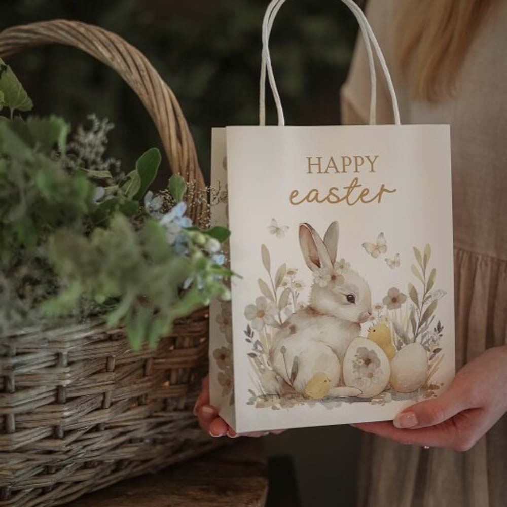 Person hält eine „Majas Cottage – Geschenketüte Frohe Ostern“ mit einem Hasenmotiv neben einem Weidenkorb gefüllt mit grünem Laub.