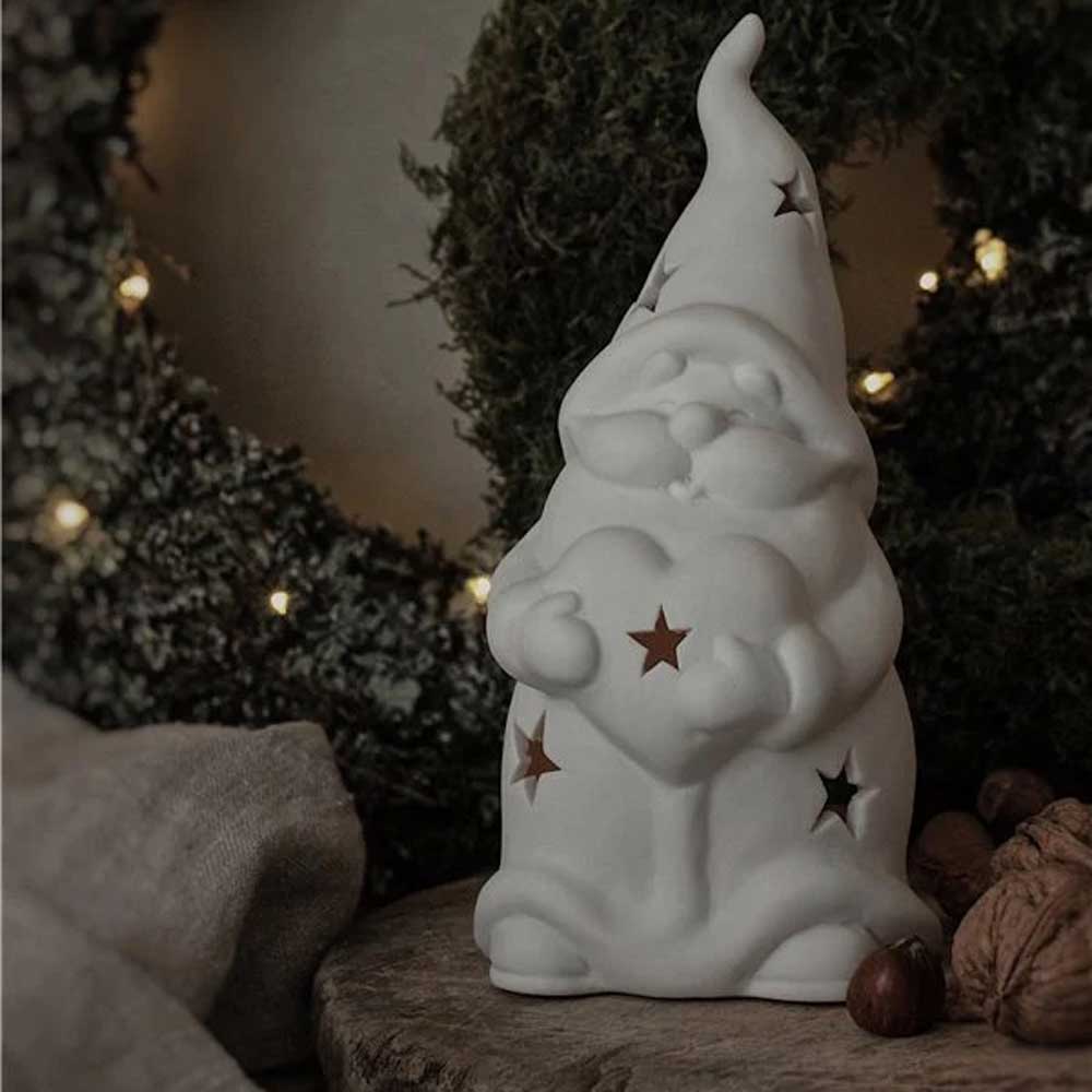 Der Majas Cottage - Mr. Santa mit Herz, eine Zwergenfigur aus Keramik mit ausgeschnittenen Sternen und einem Herz in der Hand, steht auf einer Holzfläche. Im Hintergrund ist ein Kranz mit kleinen Lichtern zu sehen.