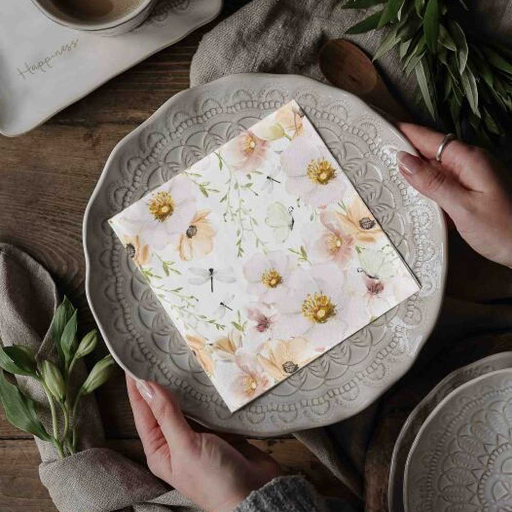 Ein Paar Hände hält einen dekorativen Teller mit einem komplizierten Muster und Majas Cottage - Papierservietten Blumen & Schmetterlinge darauf. Ein Holztisch mit Besteck, einer Tasse und Grünpflanzen umgibt das Arrangement.