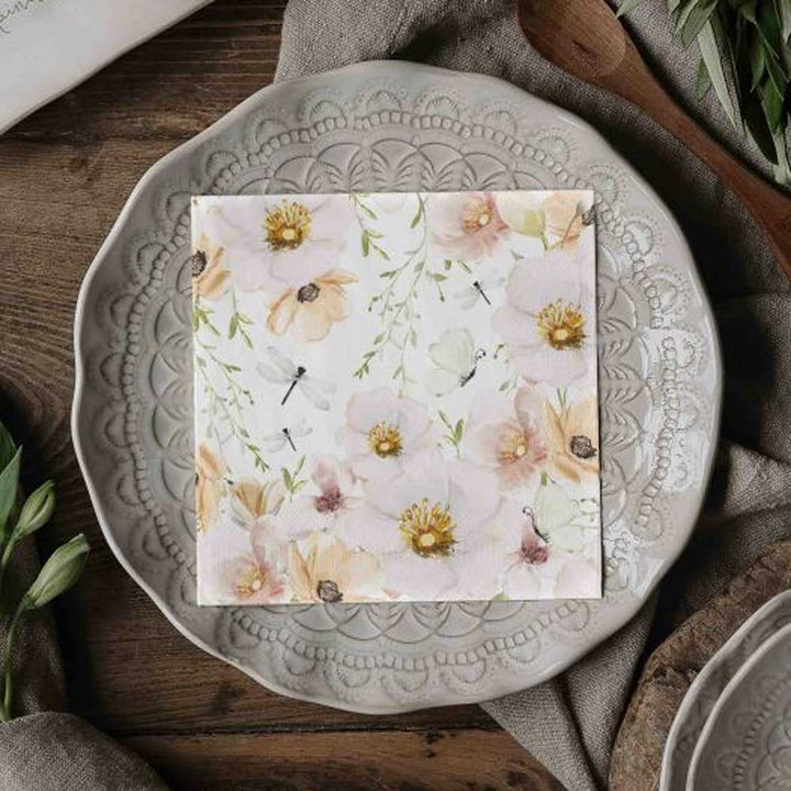 Die Papierservietten „Blumen & Schmetterlinge“ von Majas Cottage sind ordentlich gefaltet und auf einem dekorativen grauen Teller mit spitzenartigem Muster auf einem Holztisch platziert.