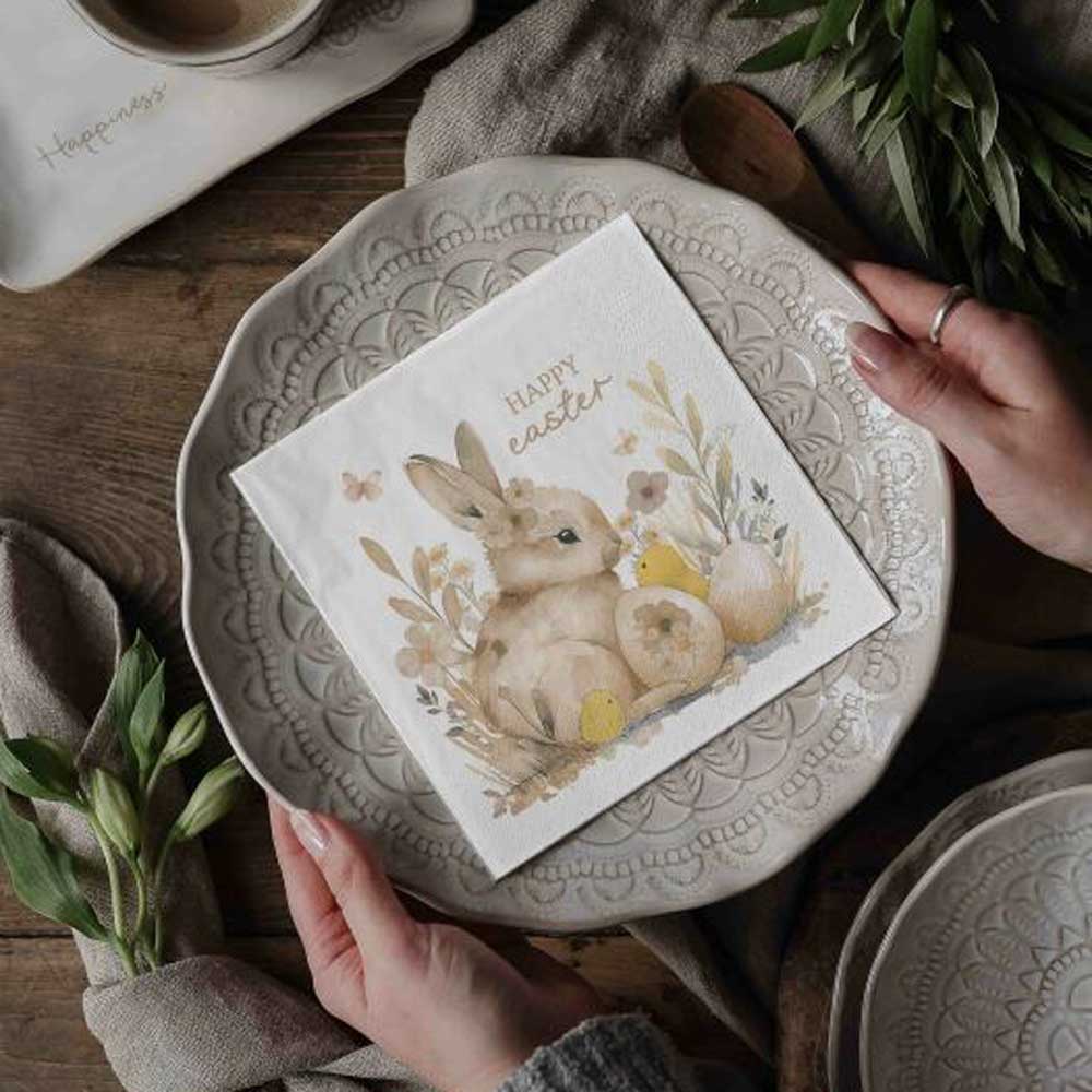 Auf einem dekorativen Teller steht „Majas Cottage - Papierservietten Frohe Ostern“, und auf der Serviette ist ein Häschen mit der Aufschrift „Frohe Ostern“ abgebildet, gedeckt auf einem rustikalen Tisch, umgeben von Grün.