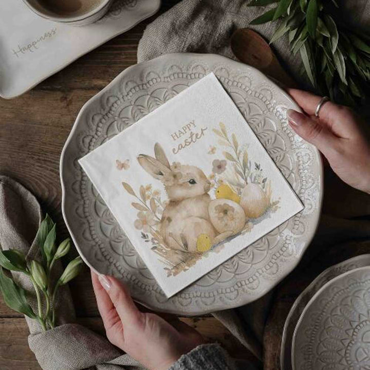 Auf einem dekorativen Teller steht „Majas Cottage - Papierservietten Frohe Ostern“, und auf der Serviette ist ein Häschen mit der Aufschrift „Frohe Ostern“ abgebildet, gedeckt auf einem rustikalen Tisch, umgeben von Grün.