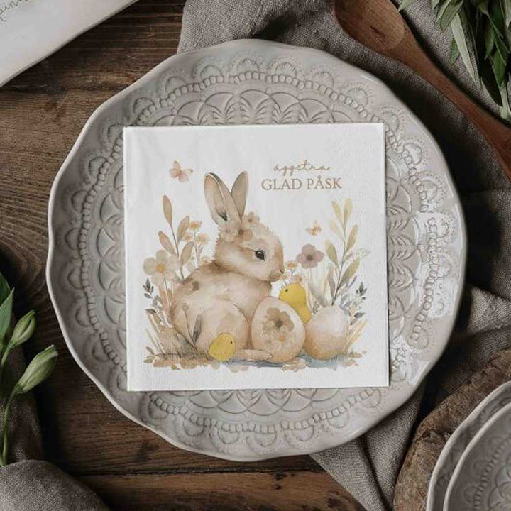 Eine Majas Cottage - Papierservietten-Serviette „Frohe Ostern“ mit einem Hasen, Blumen und Eiern liegt auf einem dekorativen Teller mit dem Text „GLAD PÅSK“.