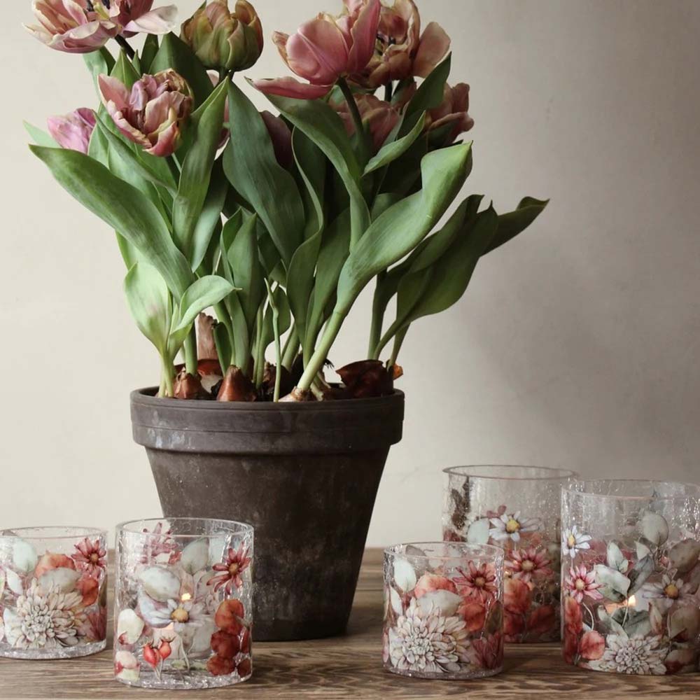 Ein schwarzer Keramiktopf mit blühenden Tulpen ist von mehreren kleinen Majas Cottage – Teelichthalter Blossom Bruchglasoptik Vintage-Glashaltern mit floralen Motiven umgeben, die alle auf einer Holzoberfläche angeordnet sind.