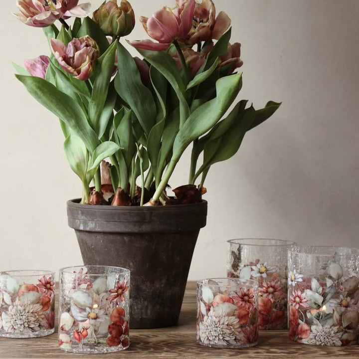 Ein schwarzer Keramiktopf mit blühenden Tulpen ist von mehreren kleinen Majas Cottage – Teelichthalter Blossom Bruchglasoptik Vintage-Glashaltern mit floralen Motiven umgeben, die alle auf einer Holzoberfläche angeordnet sind.